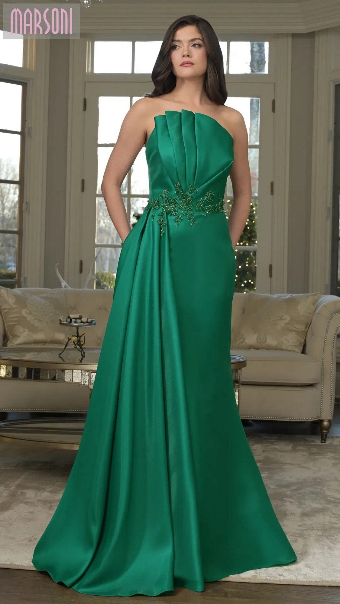 A woman in the Marsoni MV1356 long formal dress in emerald, featuring a strapless pleated bodice and floral embroidery, stands in a living room with neutral décor.